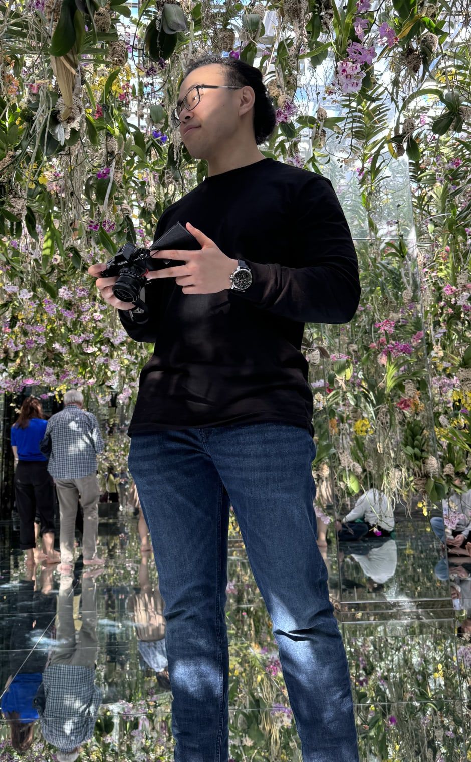 Braden Koh with a camera, standing among hanging orchids in a mirrored room.